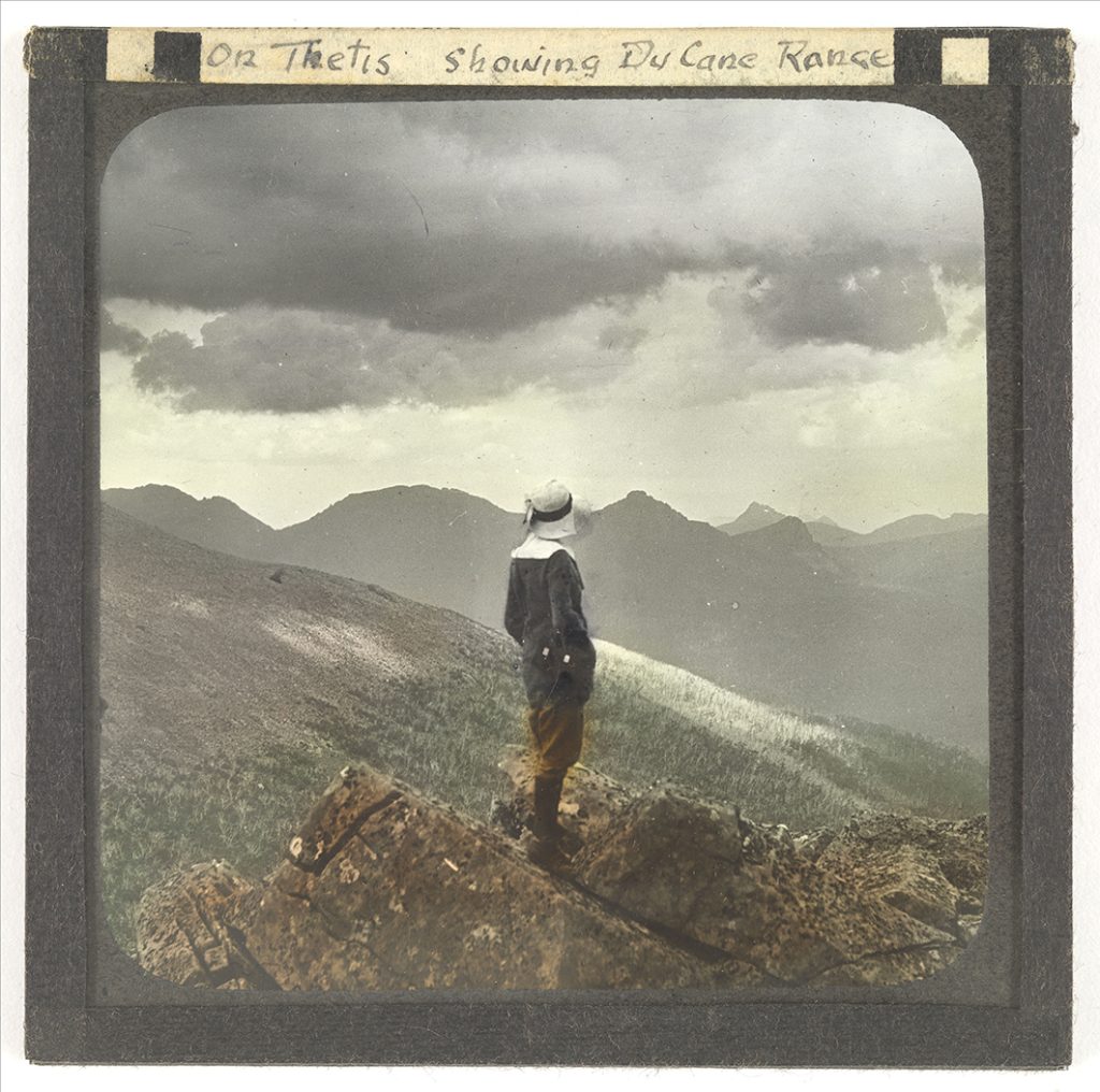 Photograph - On Thetis showing Du Cane Ranbfe [Female bushwalker standing on clifftop, watching sky and mountain tops in distance] [Lantern Slide] [Box 2]