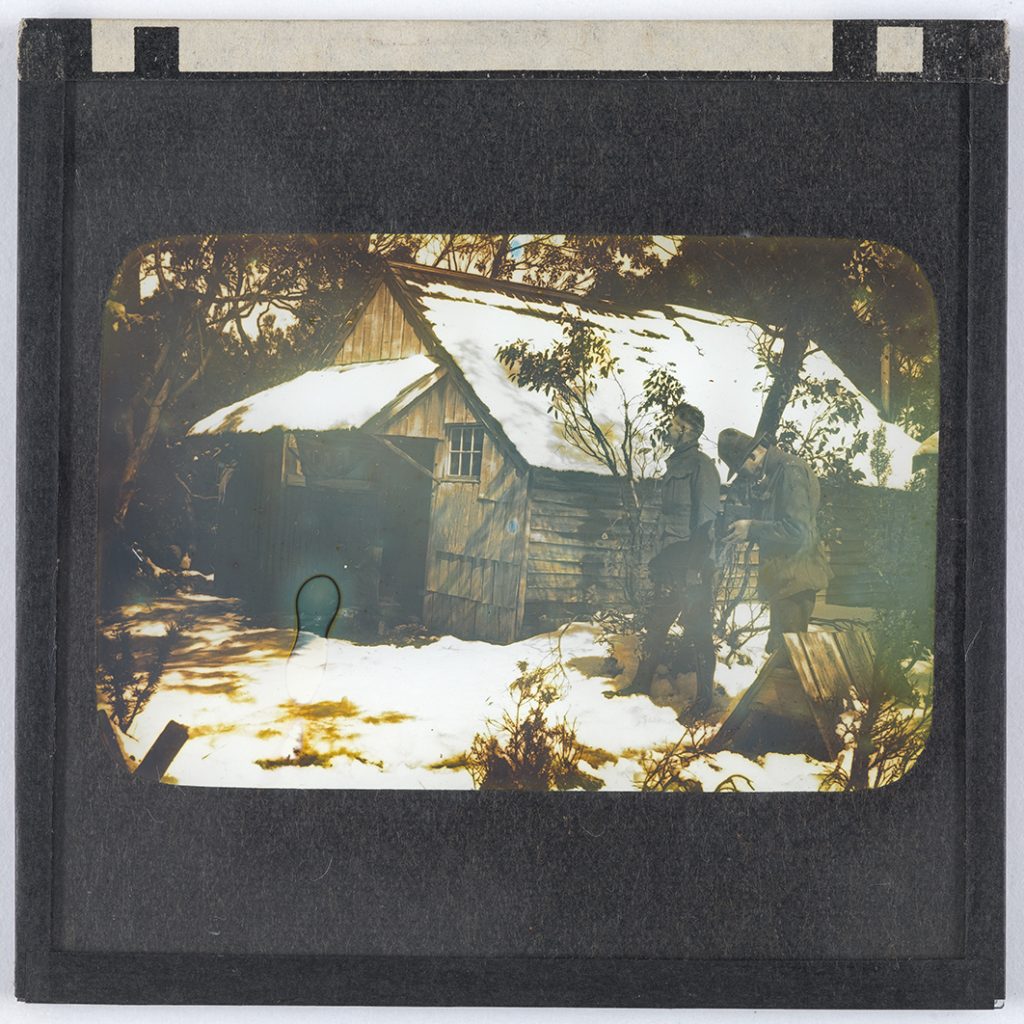 Photograph - Two men standing to side of hut (one looking down at map) [Lantern Slide - yellow tinged]