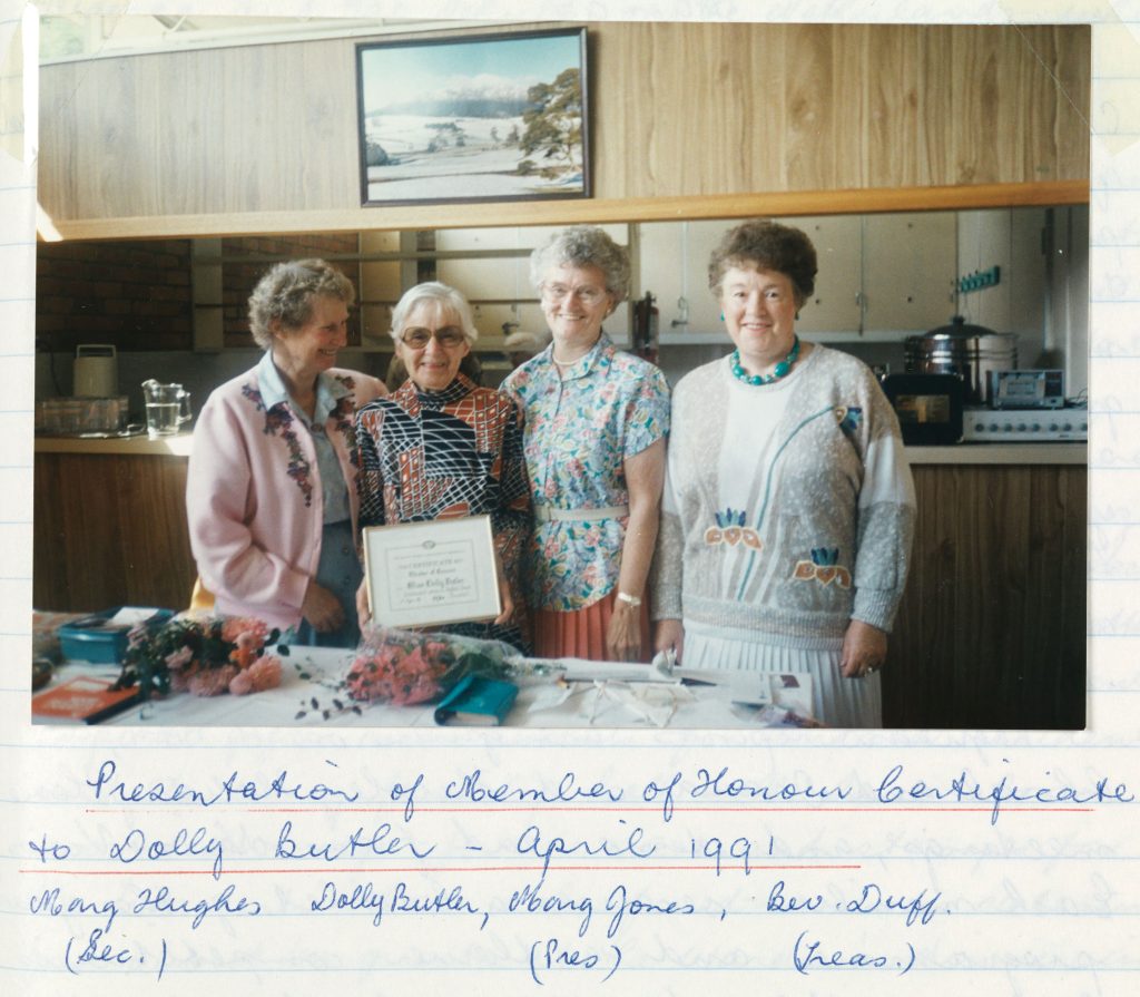 Group of four older women in a kitchen with one holding a framed certificate. Handwritten text below the photograph says 'Presentation of Member of Honour certificate to Dolly Butler April 1991. Mona Hughes (sec), Dolly Butler, Marg Jones (pres), Bev Duff (Treas).