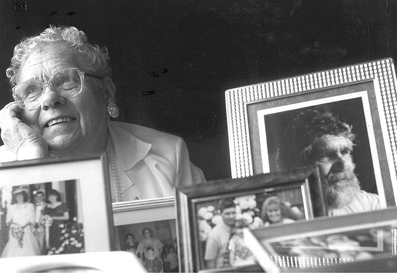 Image of Aunty Ida West, an elderly aboriginal woman with glasses standing behind several photo frames.