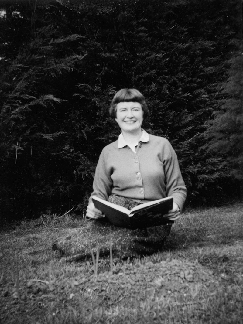 Image of Gwen Harwood smiling while sitting on the grass with an open book in her hands.