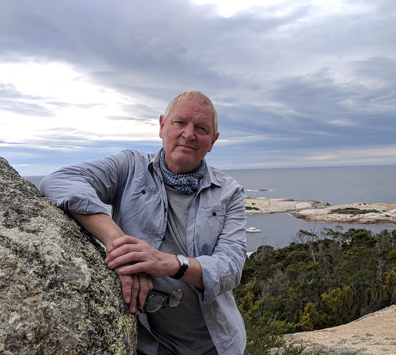 Image of Roger Scholes facing the camera standing against a rock overlooking the ocean
