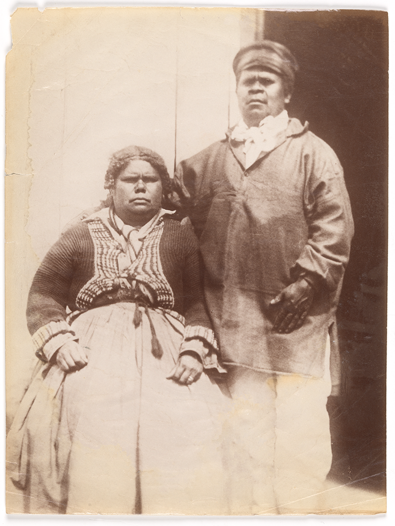 Image of Mary Ann Arthur (Clarke) and Walter George Arthur at Putalina/Oyster Cove. Two Indigenous people, a seated woman in a patterned blouse and long skirt, and a standing man in a loose shirt and hat.