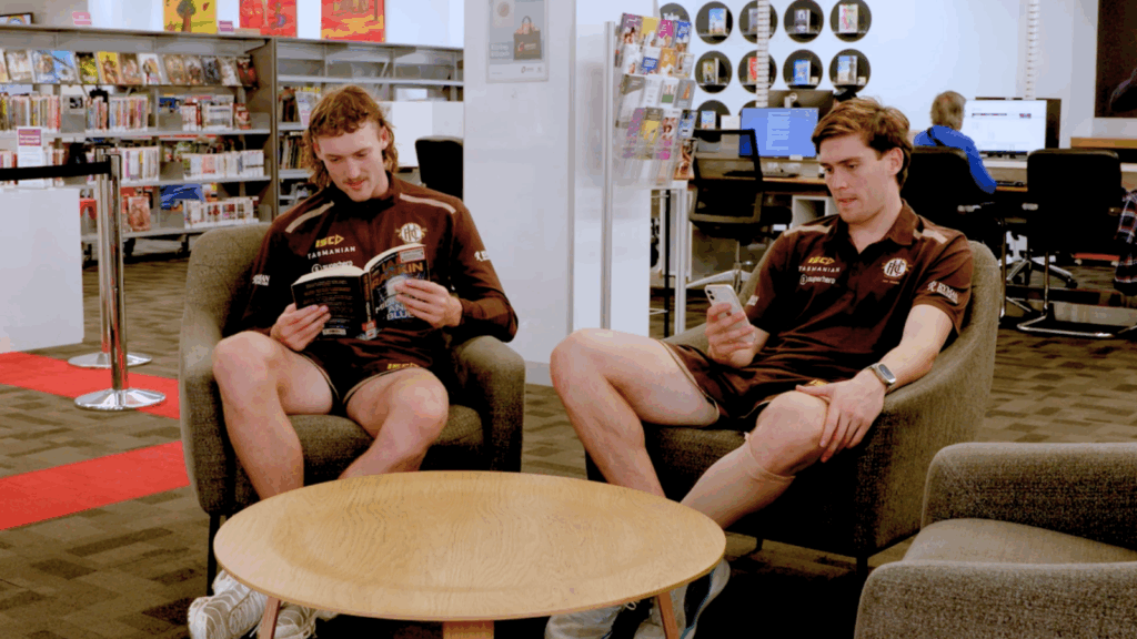 Two hawthorn football players sitting at the library. One on their phone, another reading a book.