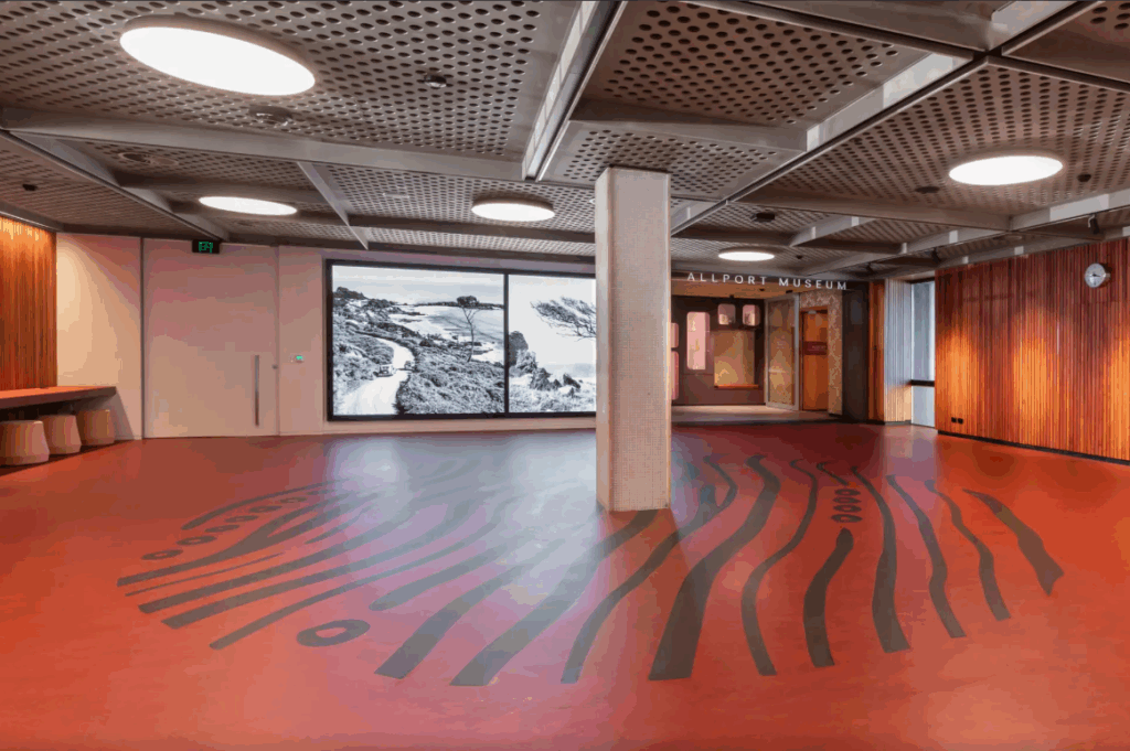 The Hobart Library ground floor. A black circular floor pattern made of lines surrounds a pillar with a big presentation screen in the background. The ceiling is grey metal with grated holes. The rest of the floor is orange lino.