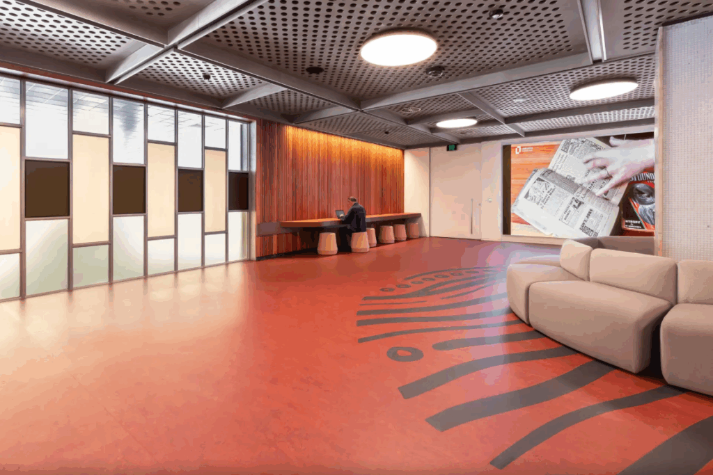 The Hobart Library ground floor. A black circular floor pattern made of lines surrounds couches that surround a pillar. The ceiling is grey metal with grated holes. The rest of the floor is orange lino.
