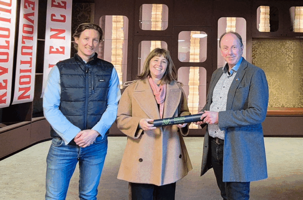 A man in a vest and jeans (Guy Edwards). A woman in a beige coat (Peta Heffernan) holding a rolled up poster with a man in a suit and patterned shirt (Ross Latham).