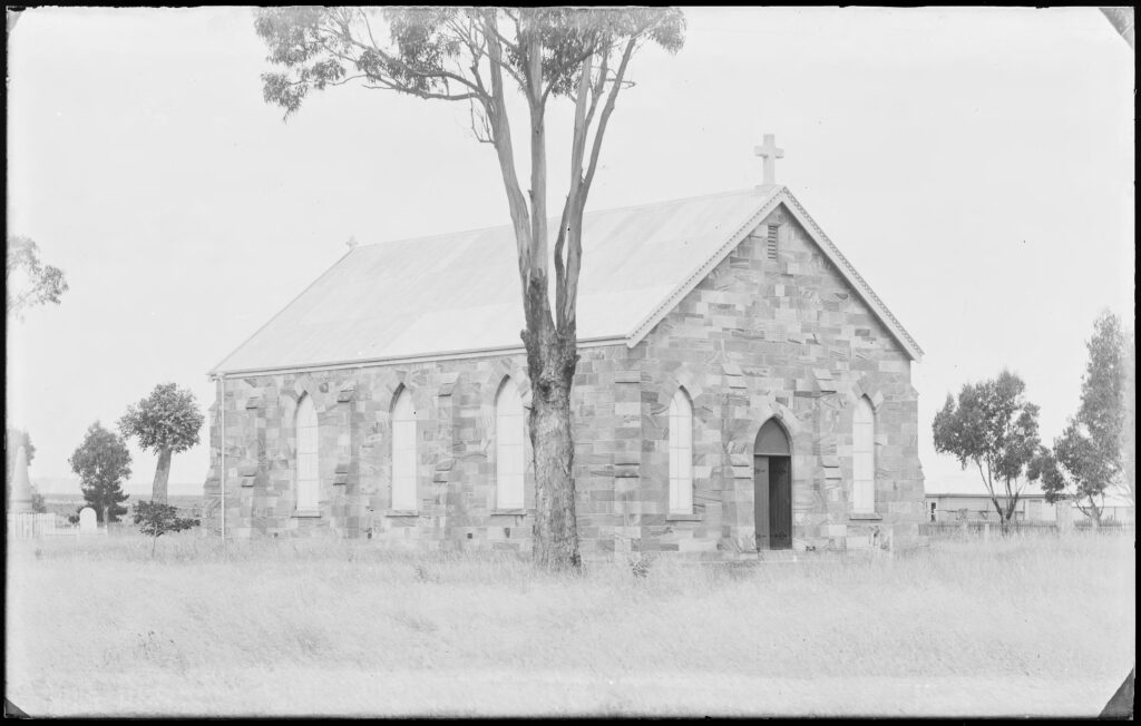 A black and white photograph of a sandstone church in a paddock with a gumtree in front