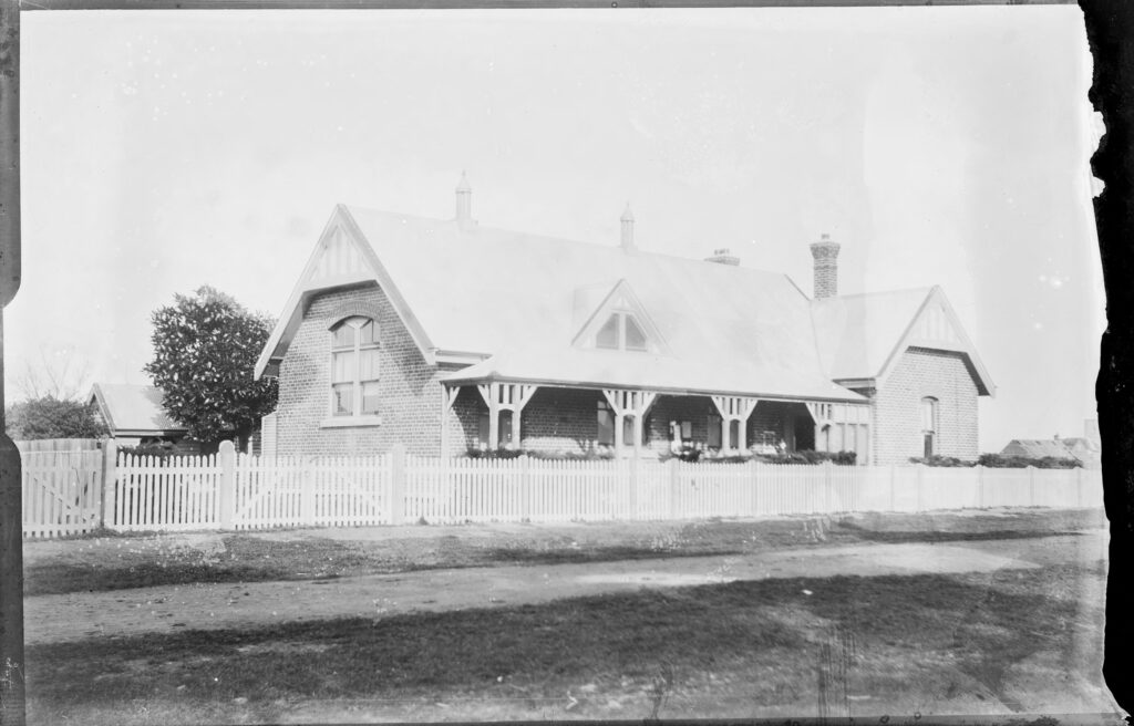 A black and white photo of a federation-era brick building with high pitched corrugated iron roof. Surrounded by a white picket fence.
