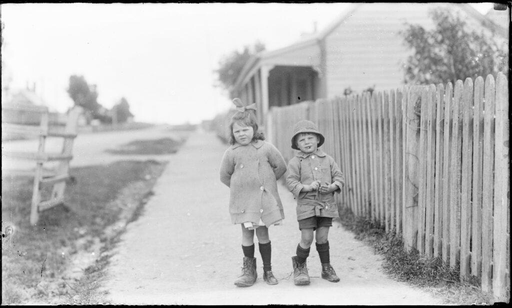 An old photo of two young children on a gravel footpath next to a fence.