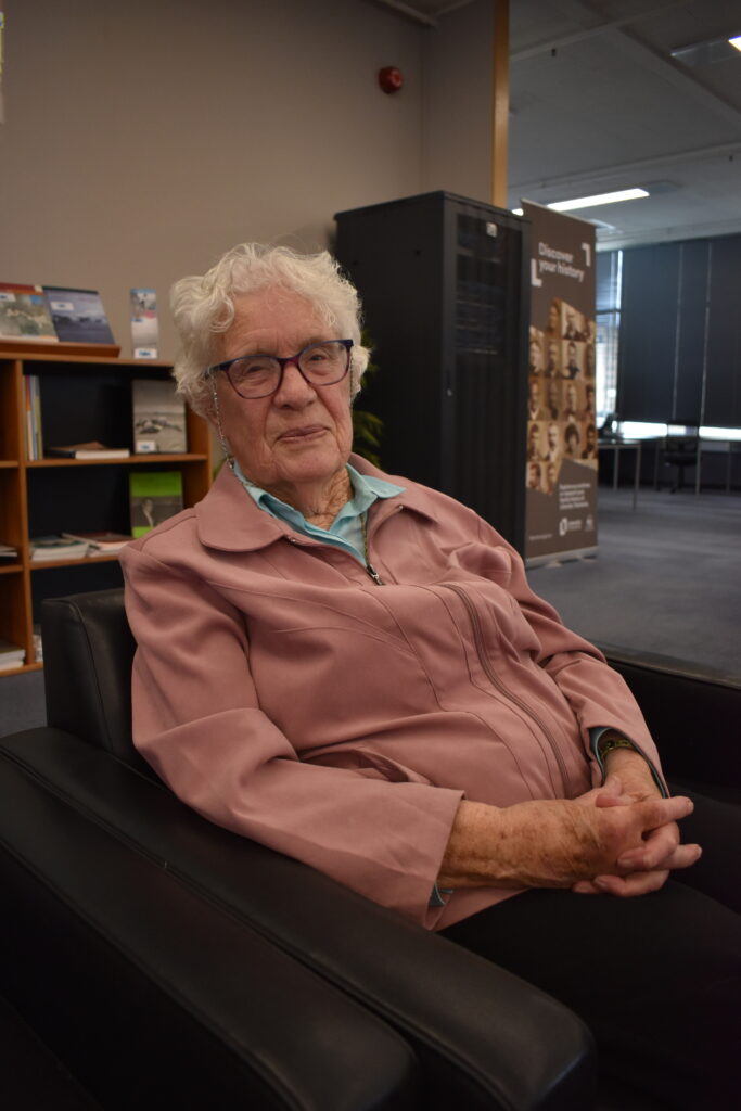Lady with short grey hair and glasses sitting in an armchair with her hands clasped in front