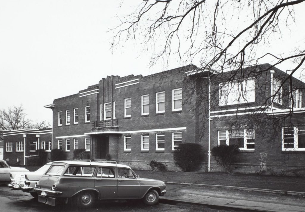 Black and white photo taken in the 1970s of the front view of an extensive two storey brick building.  Cars are parked in front of the main entrance.