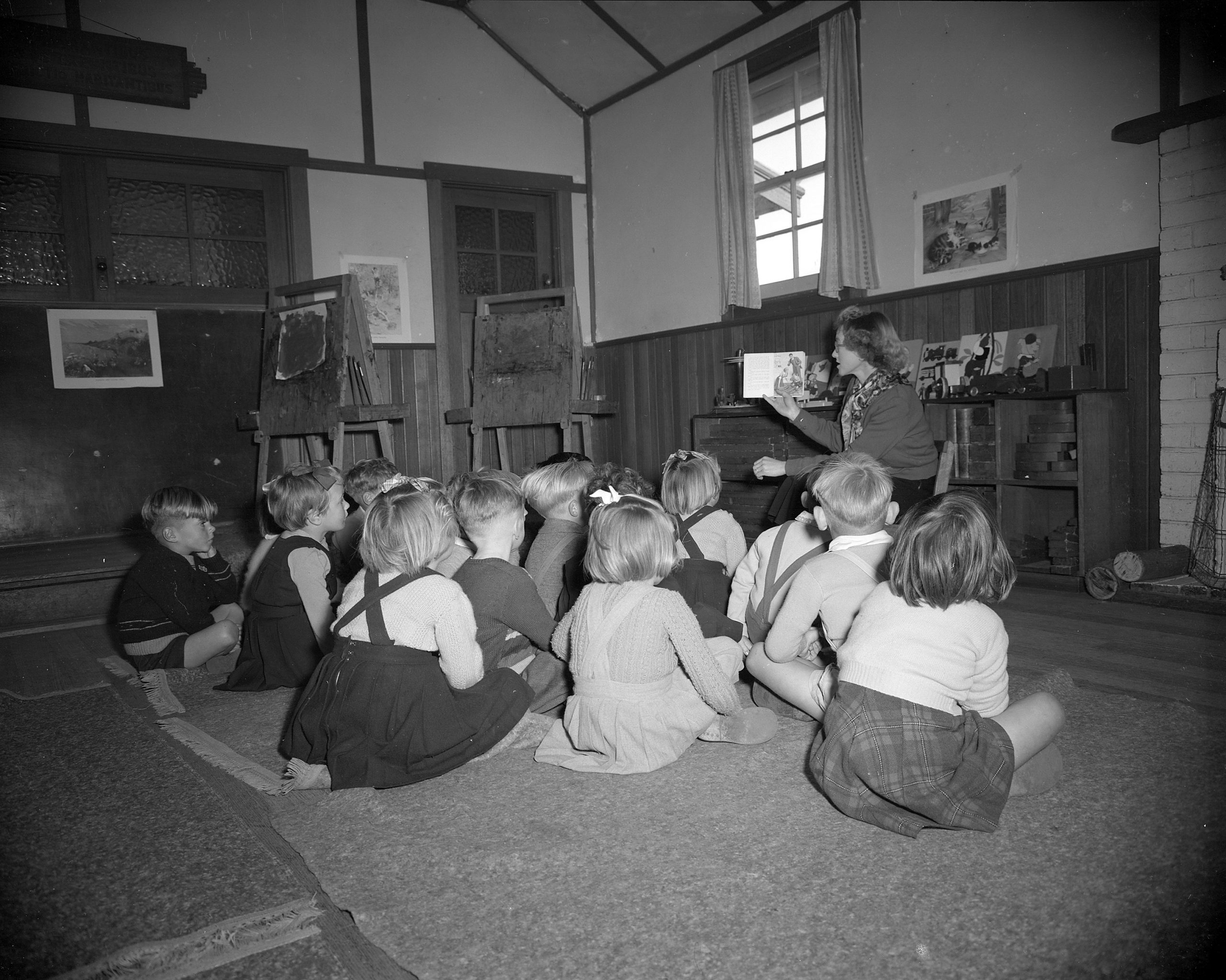 An old black and white photo of a group of school children listening to a teacher read a story inside a classroom.