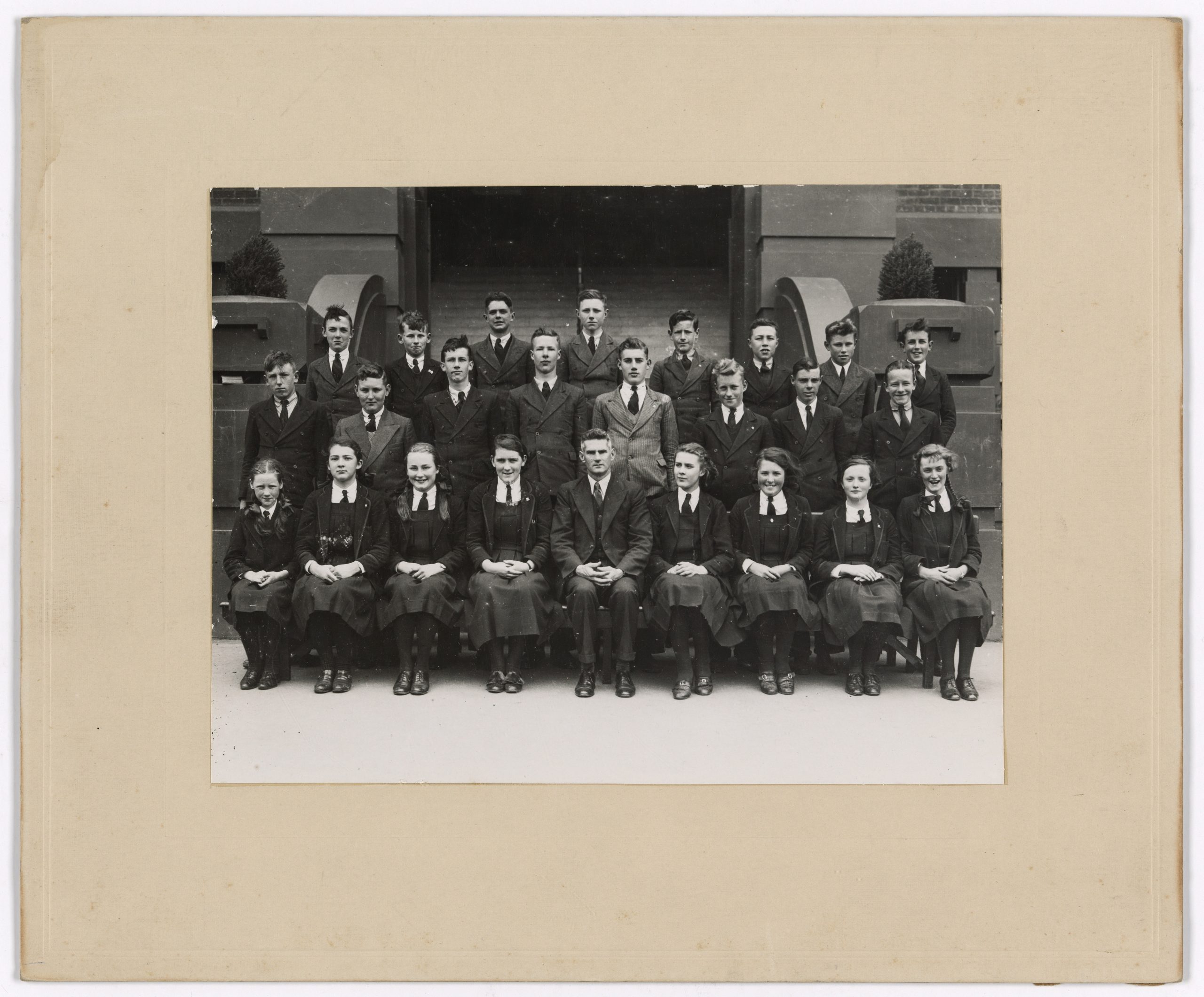 An old photo of a high school class.2 rows of boys stand behind 1 row of girls sitting, the teach sits in the front row in the middle.
