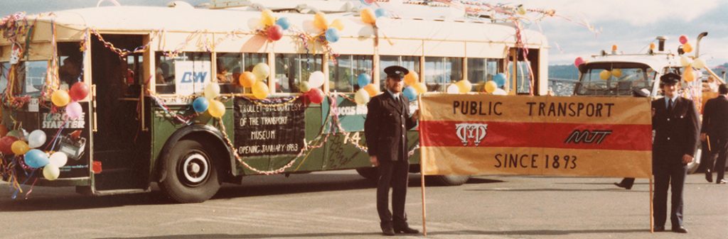 2 men holding a banner in front of a bus decorated with balloons and streamers