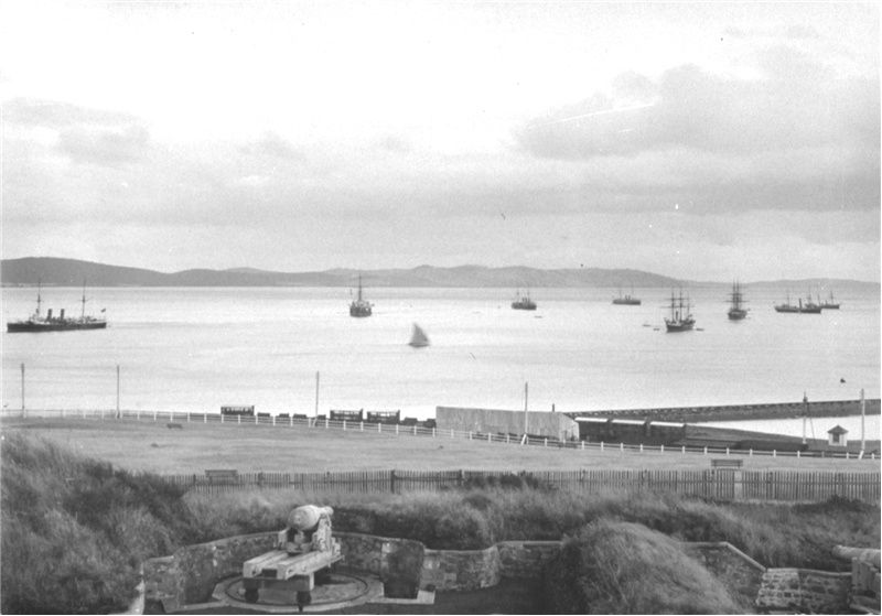 a black and white photo of ships in the ocean as seen from the shore. An artillery cannon sits on the shore in the foreground.