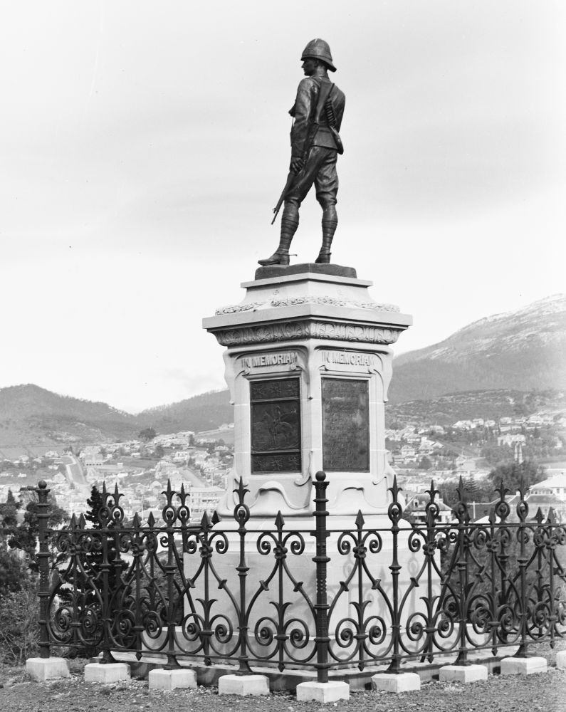 A war memorial statue. A statue of a man stand on a pillar that reads "In memoriam".  Surrounded by a fence