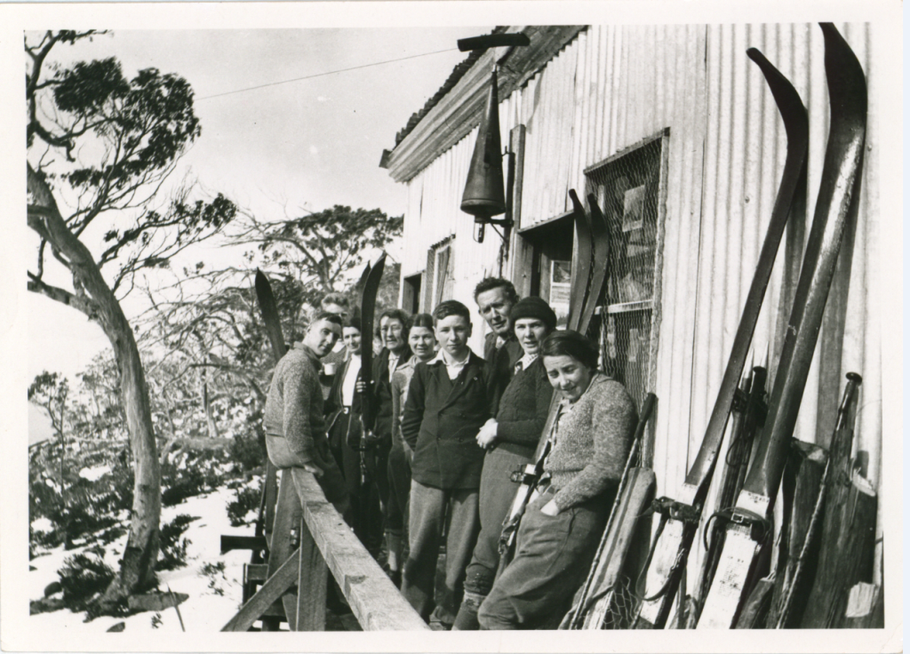 An old black and white photo of 9 people standing on the balcony of a building known as "Mulga Micks's hut." with their skis. 