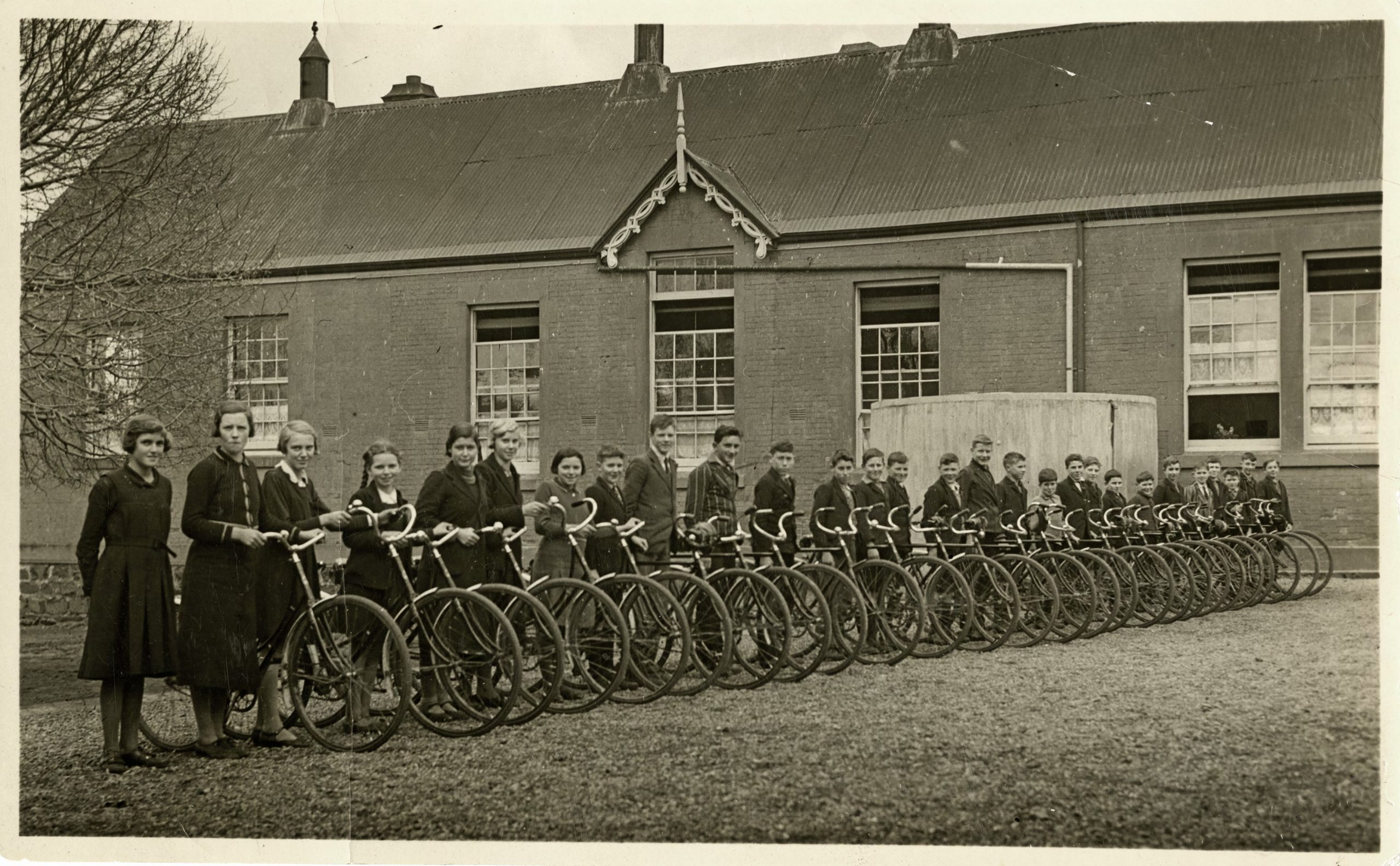 An old black and white photo of a line of school kids and their bikes.