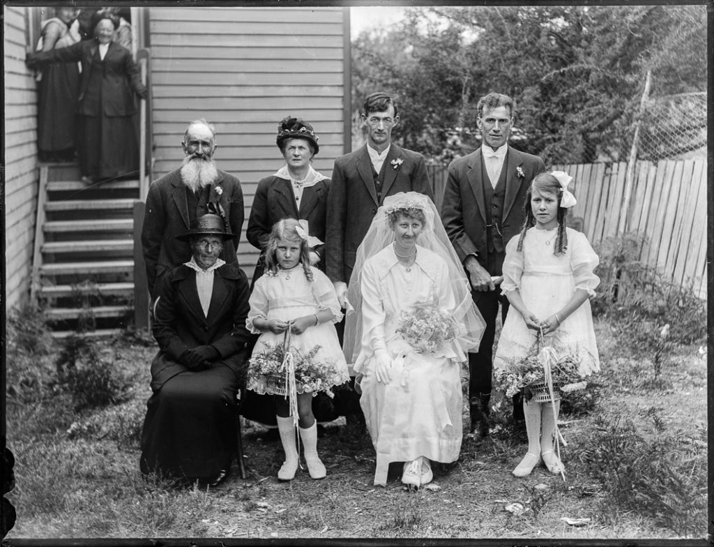 married couple in wedding outfits surrounded by family in front of the wooden house
