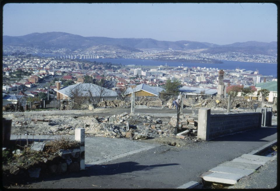 Rubble where buildings once stood. Seen from the road atop a hill. untouched houses in the distance.