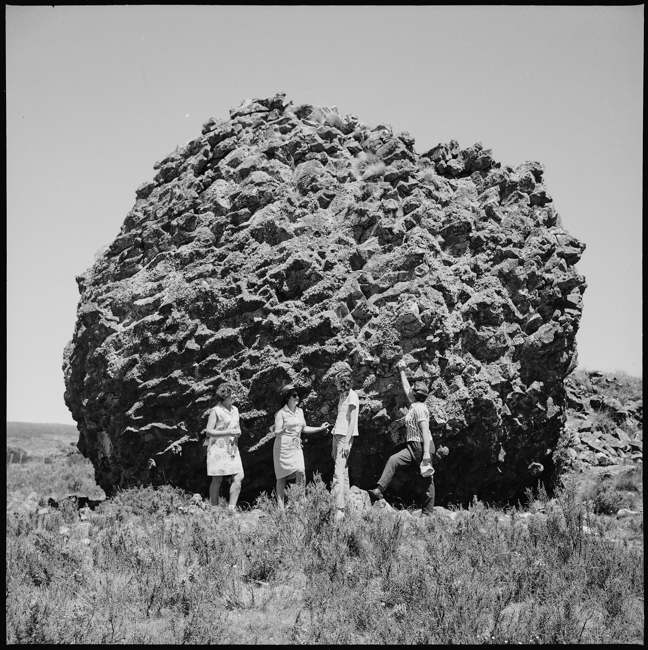 A black and white photo of 4 people standing next to a very large boulder
