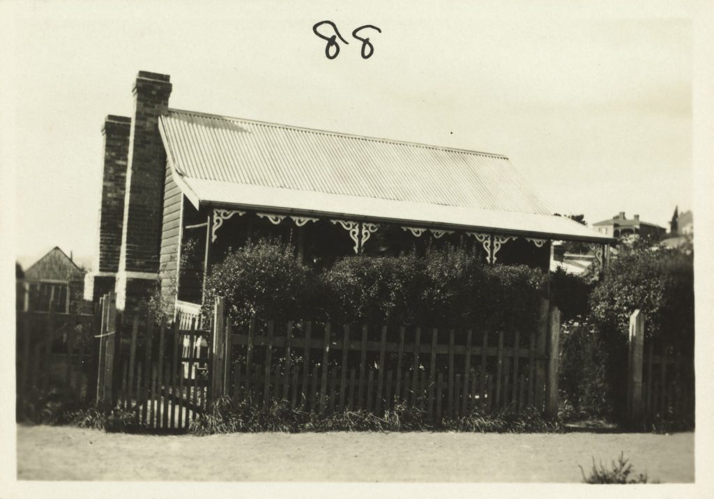 A black and white photo of a house with a picket fence and tall shrubs out front as seen from the street.