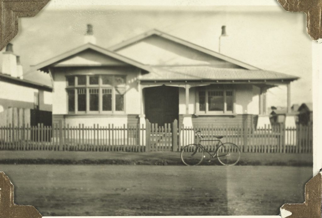 A black and white photo of a house with a fence as seen from the street. a bicycle is parked out front.