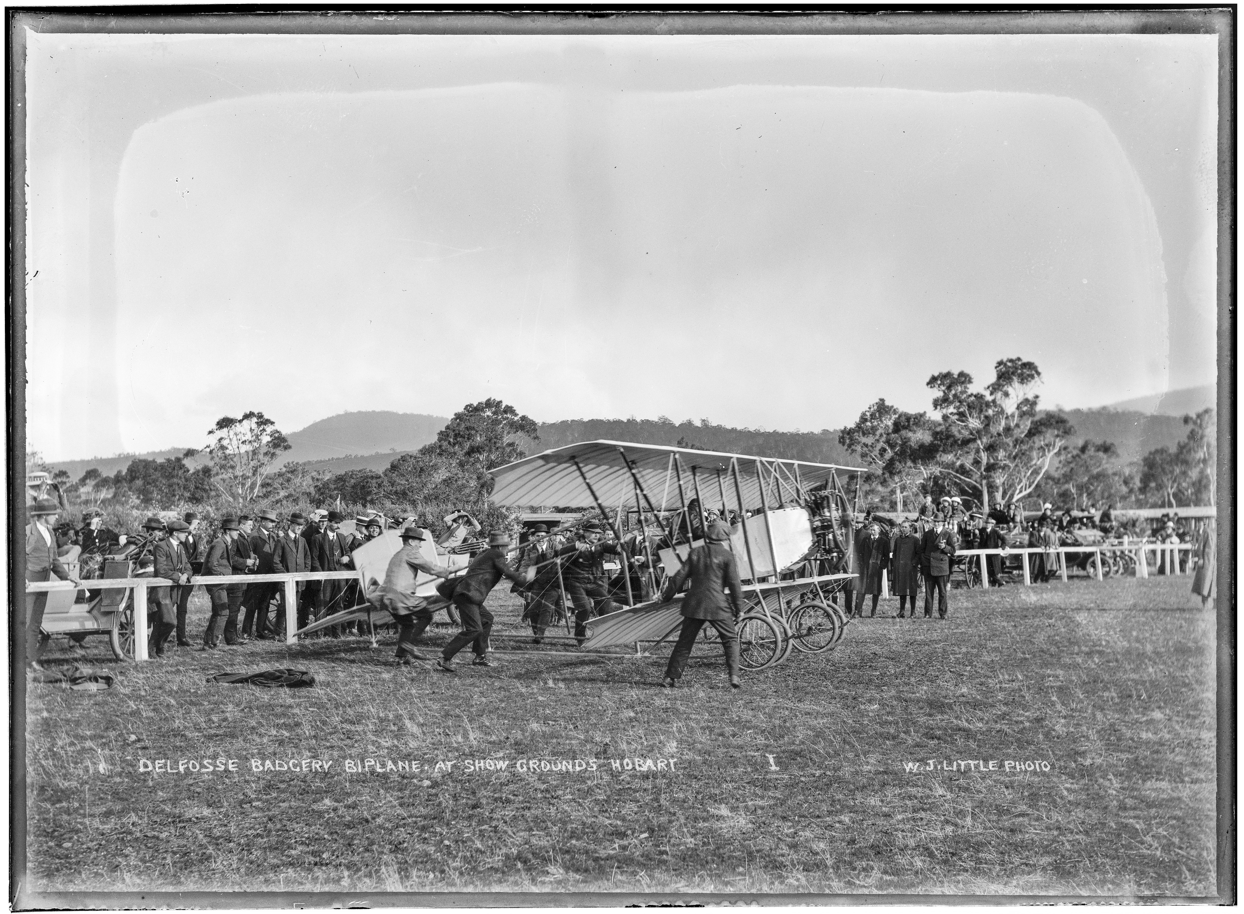 An old black and white photo of a group of people preparing a biplane for take off. a crowd of people watch.
