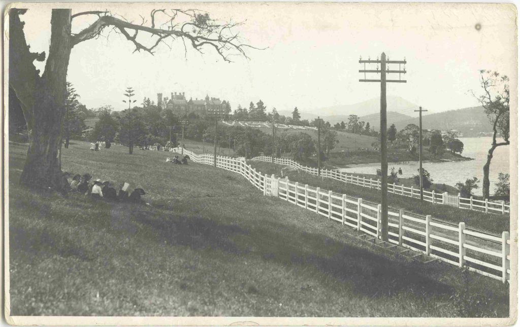 A black and white photo of a road along a grassy hill beside a river.   People picnic on the grass.  There is a large home on a hill in the background. The word "victory" has been laid out on the grass in front of the home.