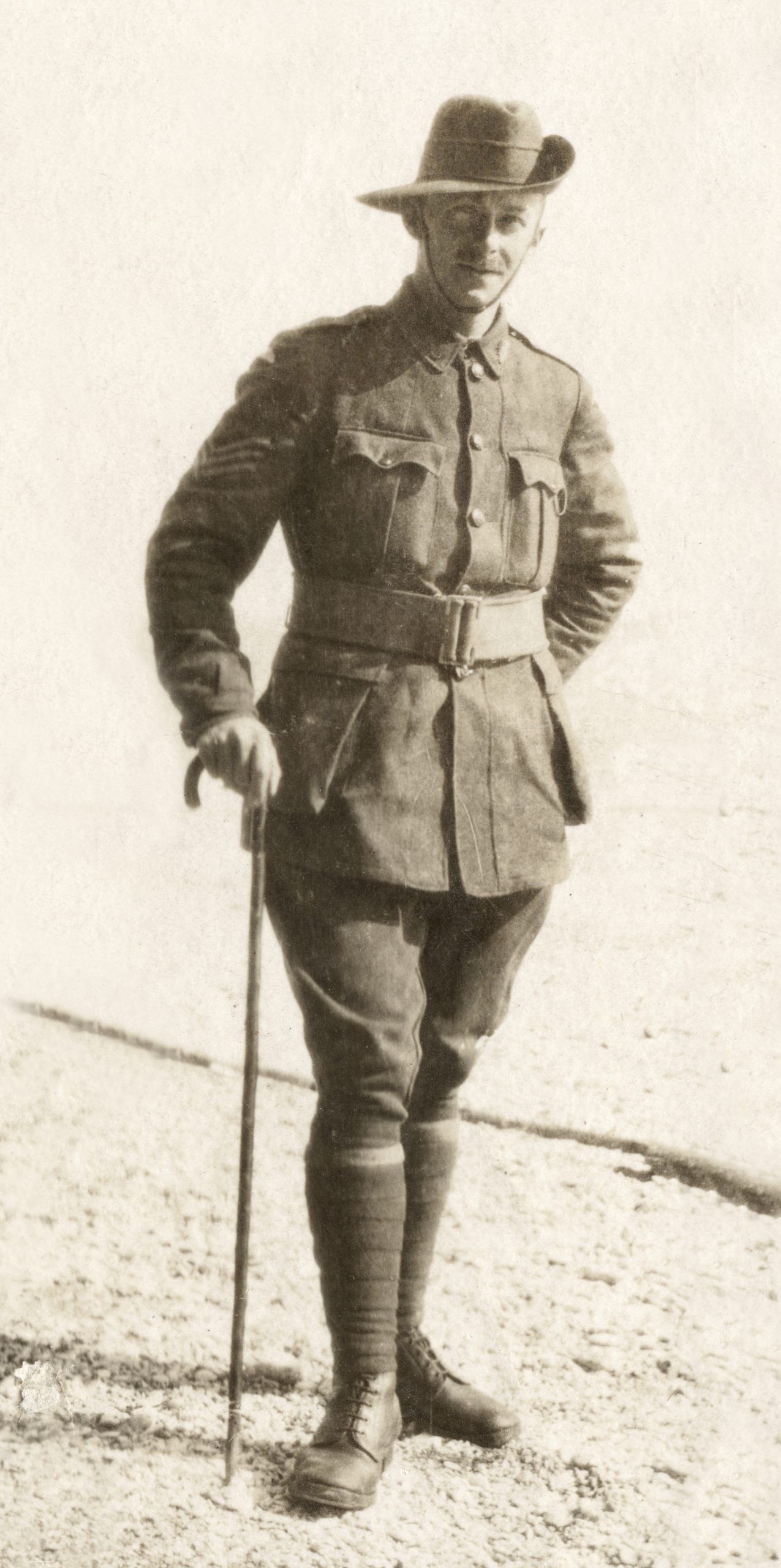 A black and white photo of a man dressed in a World War one uniform. He is standing up leaning on a walking cane held in his left hand.  