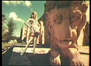a still taken from Film – The Other Face Of The Island. A woman standing next to a lion statue