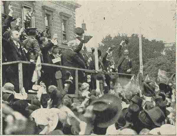 A black and white photo of a crowd of people cheering in front of a building.  Officials stand on a platform in front of the crown.