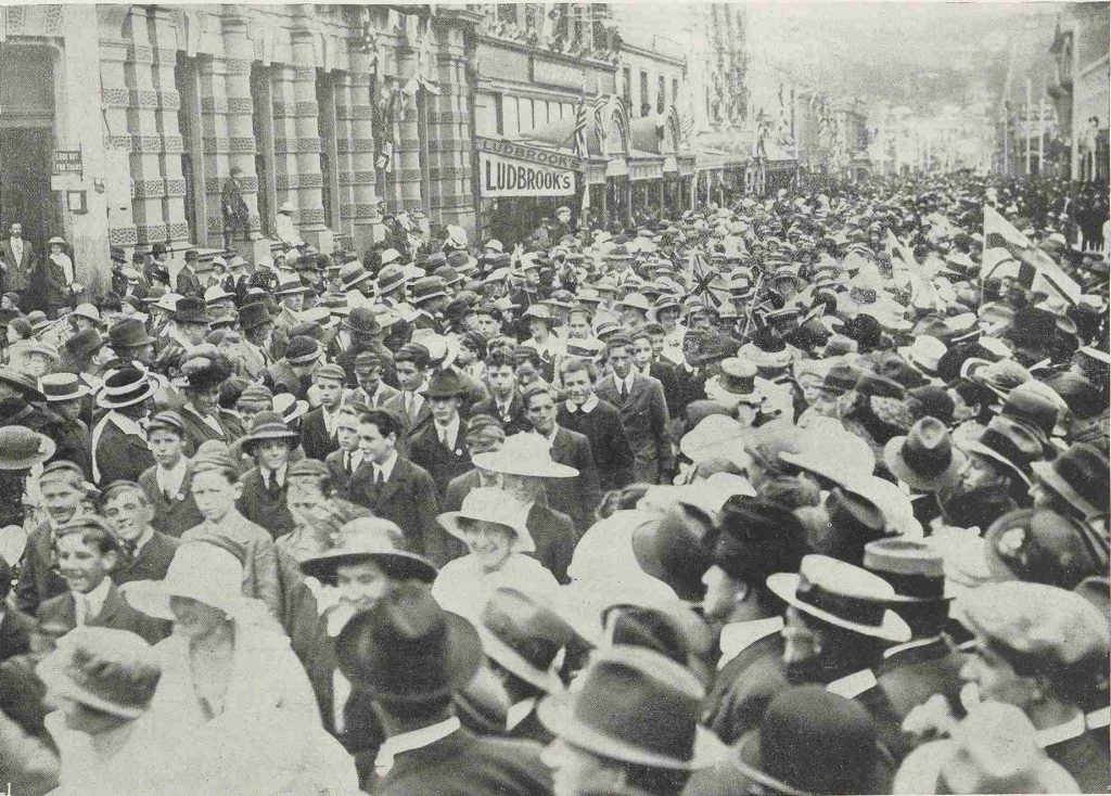 Crowd of people gather on the side of the street as a procession march along the street.