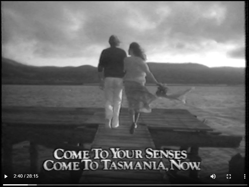 A still image from a film. 2 people walking along a pier. On sreen text is visable at the bottom. It reads: "Come to your senses come to Tasmania, now."
