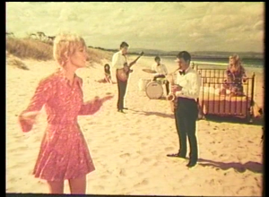 a still taken from Film – The Other Face Of The Island. 3 women on the beach with a band. one woman is on a bed